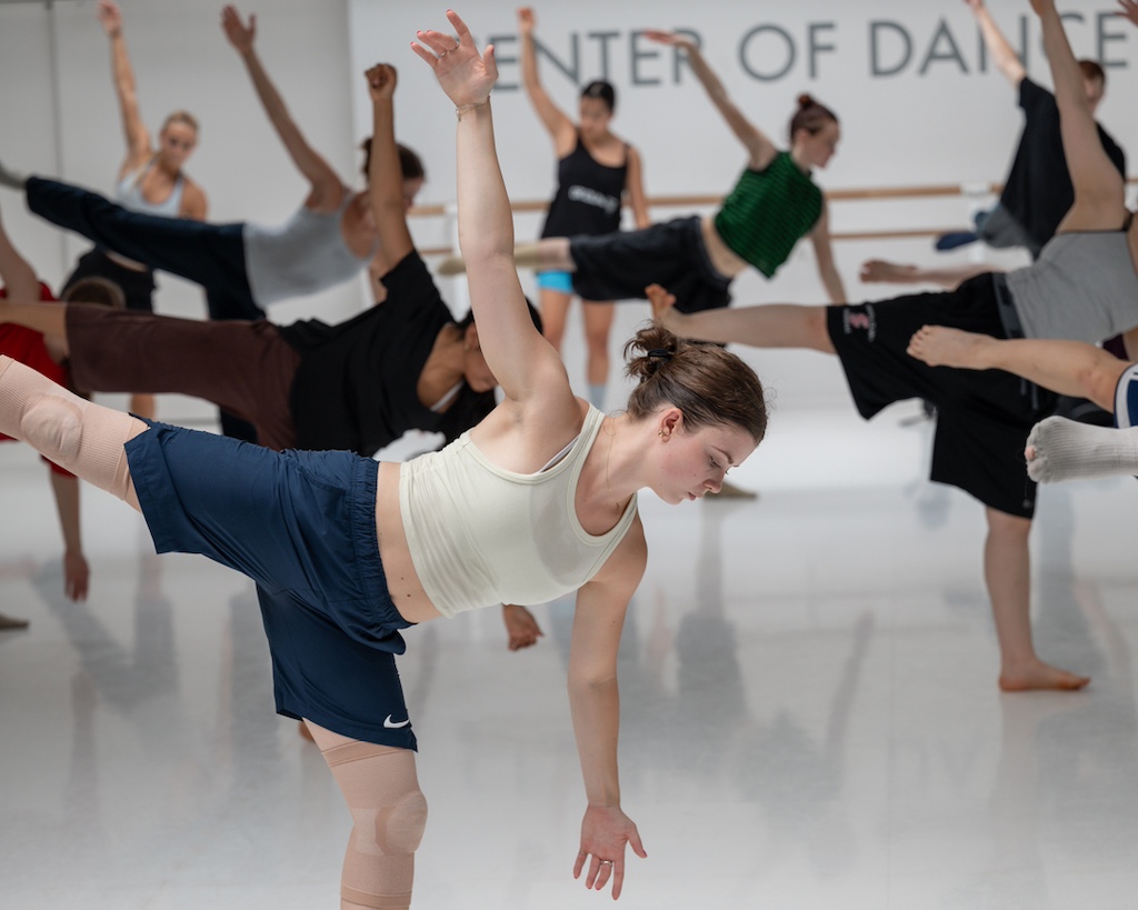 A group of dancers extend their legs while looking at the floor during a training session in a dance studio